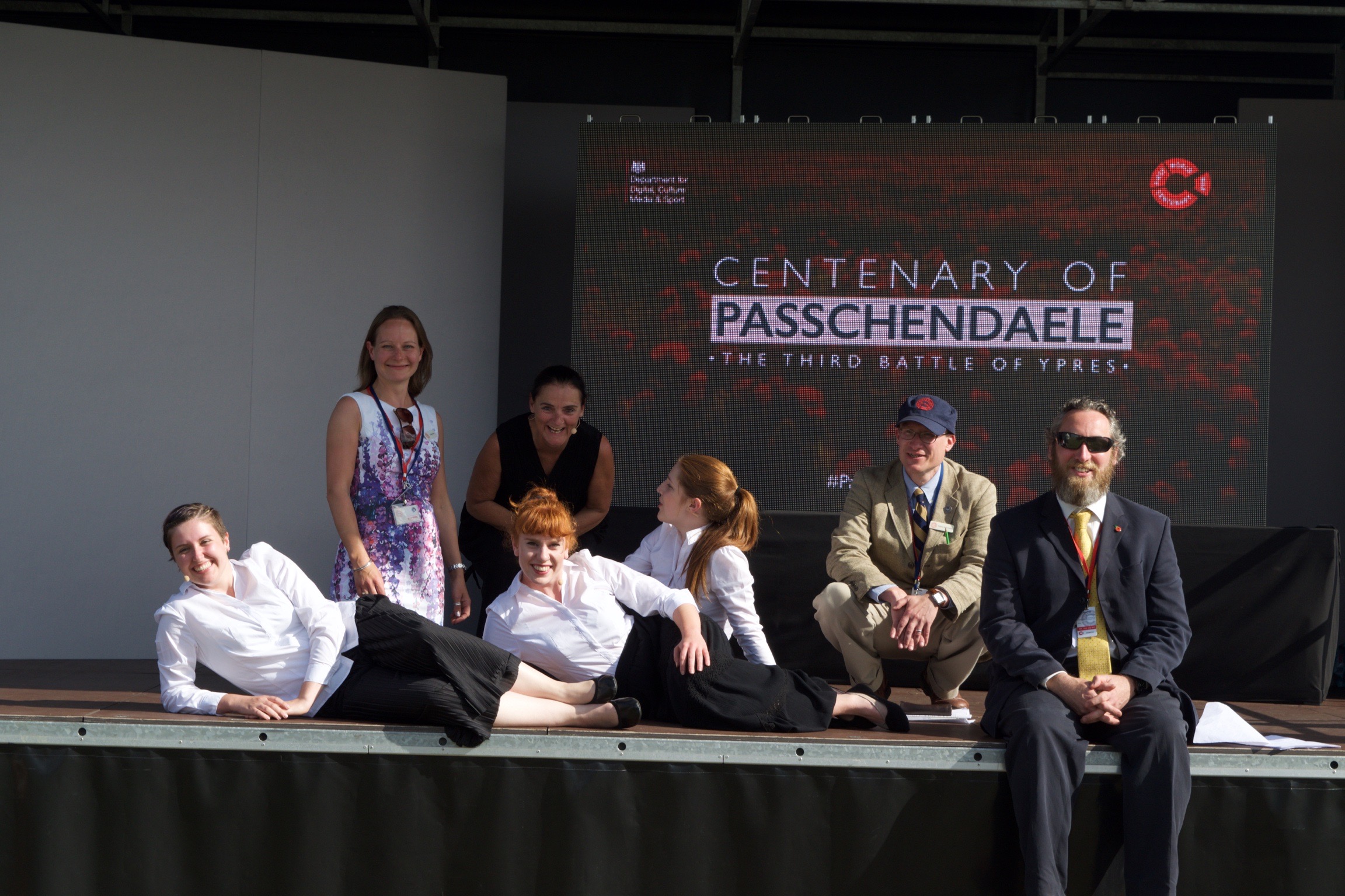 Seven members of the Gateways team posing on the outdoor stage in Belgium in front of a screen reading 'Centenary of Passchendaele, The Third Battle of Ypres'